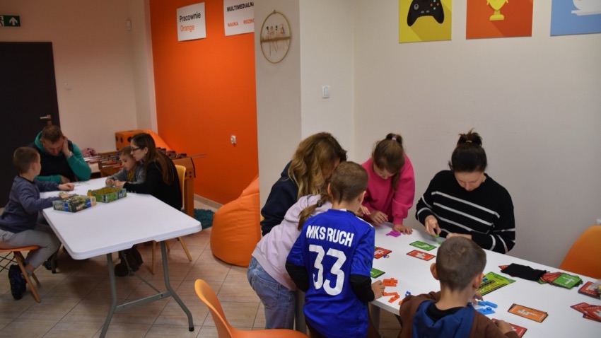 Dzień Gier Planszowych w Pracowni Orange w Wysokiem Mazowieckiem - dzieci i dorośli przy stołach z grami
